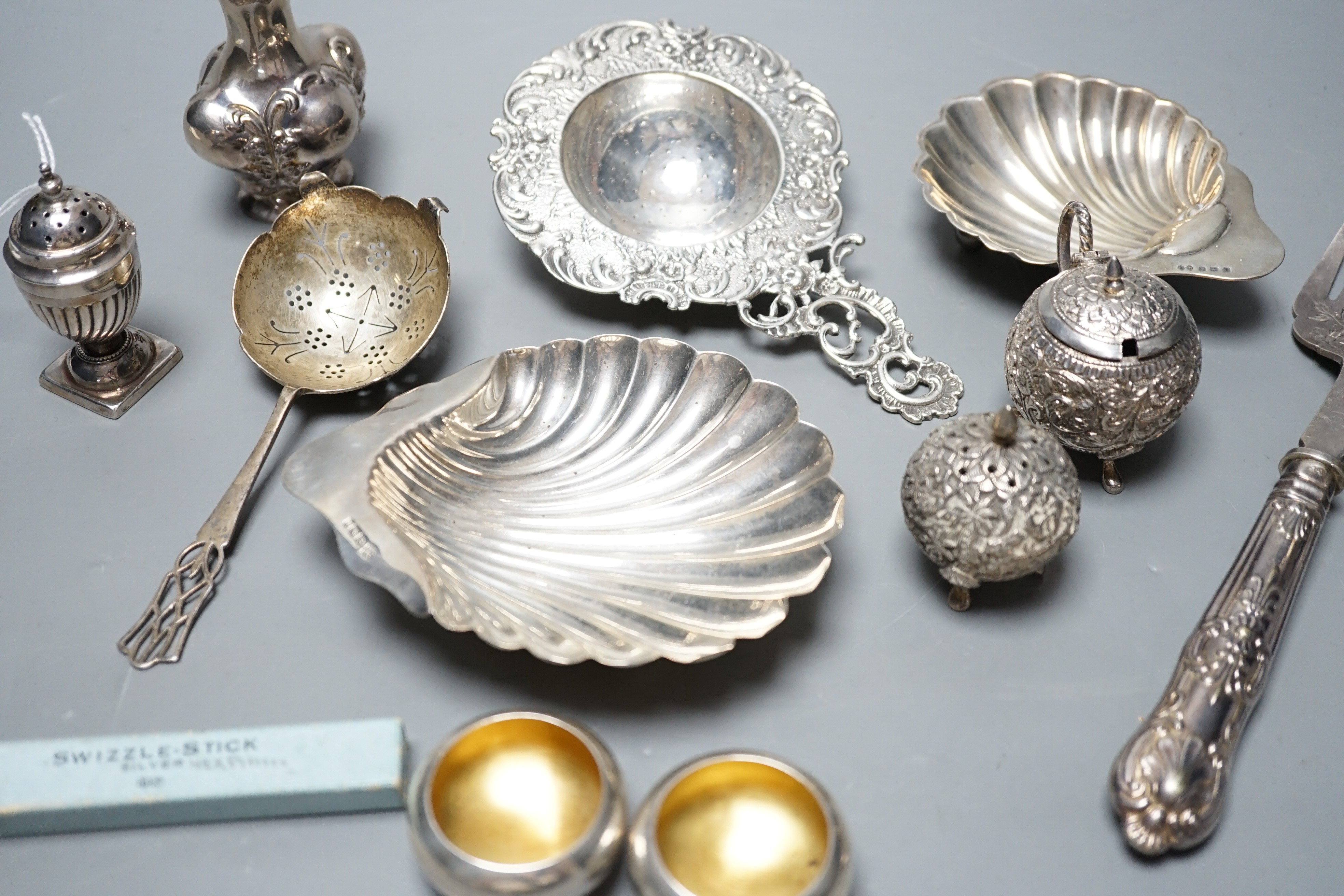 Small silver including two butter shells, a continental silver tea strainer, English tea strainer, pair of salts, swizzle stick, serving fork, two pepperettes and an Indian white metal three piece condiment set.